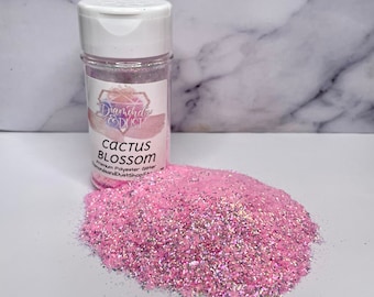 Op de afbeelding: Een fles roze glitter met het label "Cactus Blossom" staat naast een hoop van dezelfde glitter. De glitter is een mix van roze en iriserende deeltjes. De fles heeft een witte dop en een etiket met een logo en productinformatie.