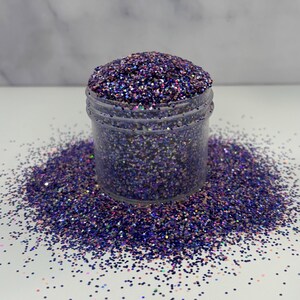 May include: A clear plastic jar filled with purple glitter, with some glitter spilled around the base. The glitter has a mix of purple, blue, and iridescent colors, creating a sparkling effect. The jar is sitting on a white surface.