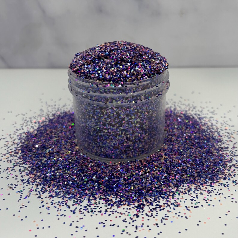 May include: A clear plastic jar filled with purple glitter, with some glitter spilled around the base. The glitter has a mix of purple, blue, and iridescent colors, creating a sparkling effect. The jar is sitting on a white surface.