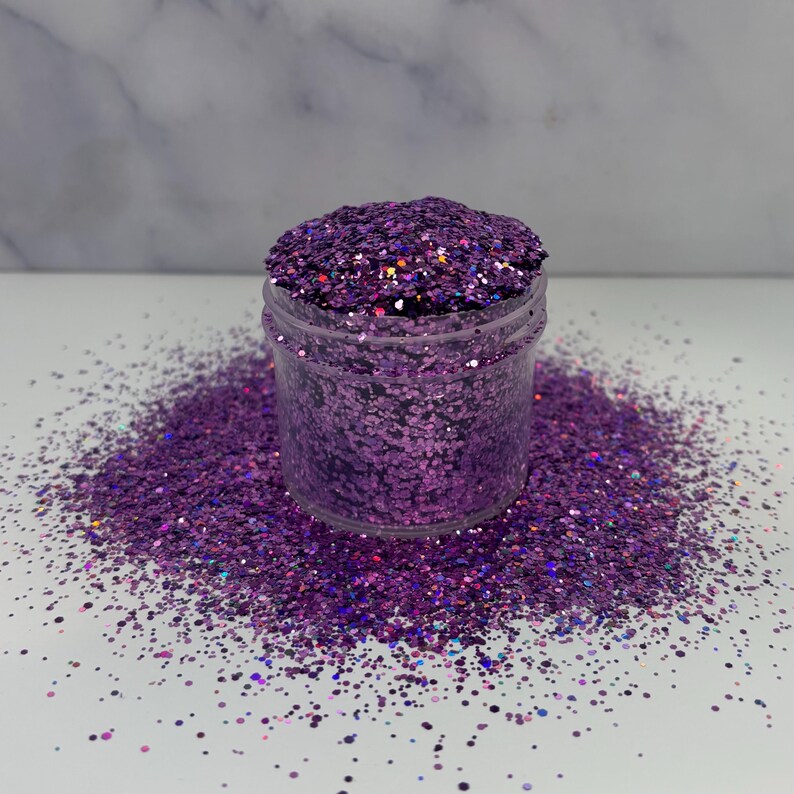 May include: A clear plastic jar filled with vibrant purple glitter, with some glitter spilled around the base. The glitter has a mix of shapes and sizes, reflecting light. The background is a white surface with a marble pattern.