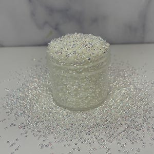 May include: A clear plastic jar filled with small, iridescent beads. The beads are scattered around the jar on a white surface. The beads have a pearlescent, shimmering quality, reflecting light. The jar is transparent, allowing the beads to be seen.