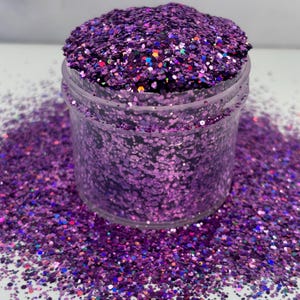 May include: A clear plastic container filled with vibrant purple glitter. The glitter is a mix of shapes and sizes, with a sparkling, iridescent quality. Some glitter has a rainbow sheen. The container sits on a surface covered in scattered glitter.