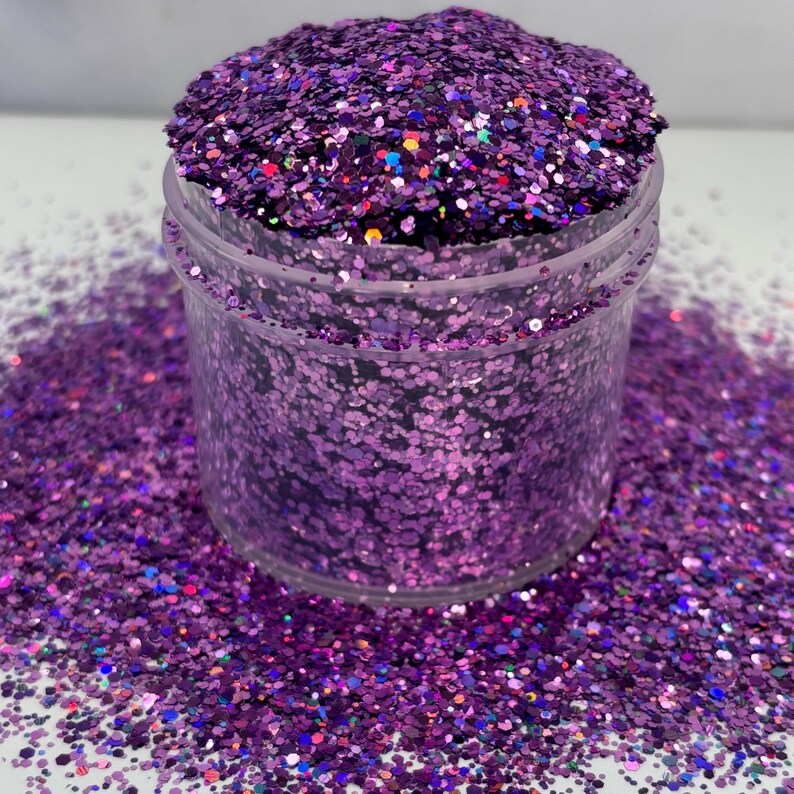 May include: A clear plastic container filled with vibrant purple glitter. The glitter is a mix of shapes and sizes, with a sparkling, iridescent quality. Some glitter has a rainbow sheen. The container sits on a surface covered in scattered glitter.
