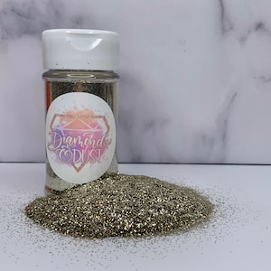 Puede incluir: Un frasco de plástico transparente lleno de brillantina dorada fina. El frasco tiene una tapa blanca y una etiqueta que dice "Diamond Dust".