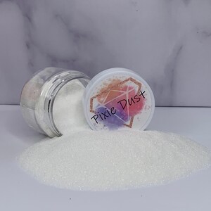 May include: A clear plastic jar tipped on its side, spilling fine white glitter onto a surface. The white lid features a diamond-shaped graphic with a watercolor-style background in shades of orange, pink, and purple, with the text Pixie Dust.