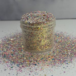 May include: A clear plastic jar filled with iridescent glitter. The glitter is a mix of colors, including pink, blue, silver, and gold. Some glitter is scattered around the jar on a white surface.