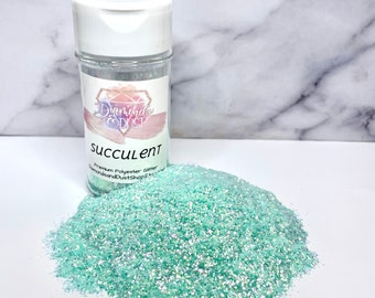 Op de afbeelding: Een doorzichtige plastic pot gevuld met lichtgroene glitter met een iriserende glans. De pot heeft een wit deksel en een etiket met de tekst "Diamond Dust" en "Succulent" in roze en witte letters. Premium Polyester Glitter.