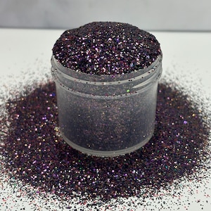 May include: A clear jar filled with iridescent purple, black, and green glitter. The glitter is scattered around the jar on a white surface.