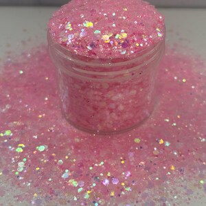 May include: A clear plastic jar filled with pink glitter. The glitter is a mix of sizes and shapes, with iridescent flecks that catch the light. Some glitter is scattered around the jar.