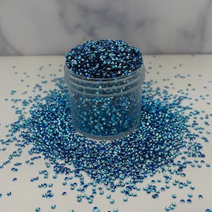 May include: A clear plastic jar filled with small, iridescent blue sequins. The sequins are scattered around the jar on a white surface. The sequins have a shiny, reflective quality, creating a sparkling effect.