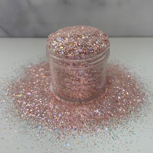 May include: A clear glass jar filled with pink and white iridescent glitter. The glitter is scattered around the jar on a white surface.