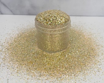 May include: A clear plastic jar filled with gold glitter, with some glitter spilled around it. The glitter has a mix of gold and silver tones, creating a sparkling effect. The jar is sitting on a white surface.