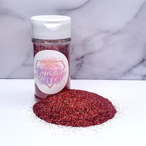 May include: A clear plastic bottle filled with red glitter, next to a pile of loose glitter. The bottle has a white lid and a label with the text "Diamonds & Dust" in a decorative font. The glitter is scattered on a white surface.