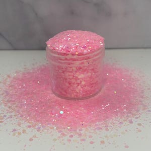 May include: A clear plastic jar filled with pink glitter, with some glitter spilled around the base. The glitter is a mix of different shapes and sizes, creating a sparkling effect. The background is a white surface.