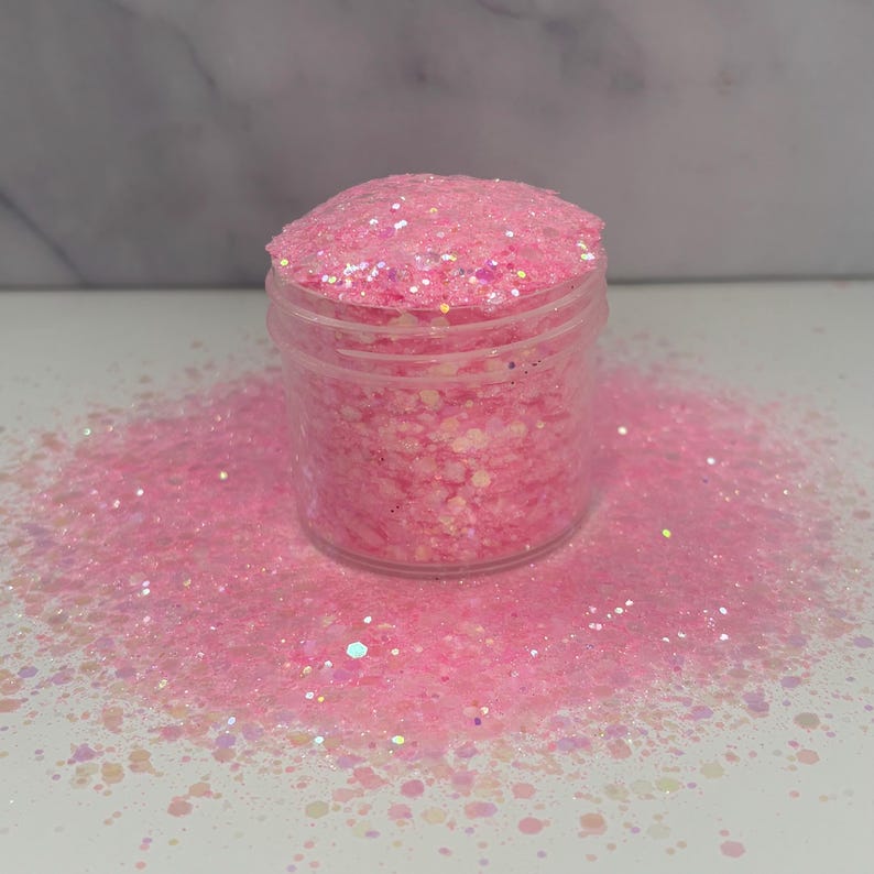 May include: A clear plastic jar filled with pink glitter, with some glitter spilled around the base. The glitter is a mix of different shapes and sizes, creating a sparkling effect. The background is a white surface.
