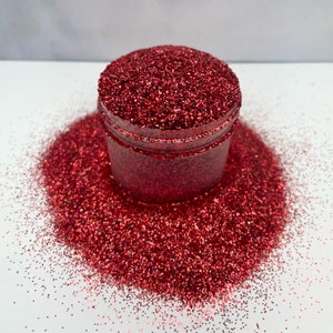 May include: A jar filled with red glitter, surrounded by a generous scattering of the same sparkling material. The glitter is a deep, rich red color, catching the light and creating a dazzling effect. The jar is clear, allowing the glitter to be fully visible.