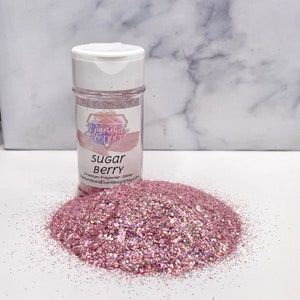 May include: A clear plastic bottle of "Sugar Berry" glitter with a white cap, next to a pile of loose glitter. The glitter is a mix of pink and iridescent particles, reflecting light. The bottle has a label with the brand name and product details.