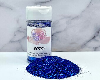 May include: A clear jar filled with blue glitter labeled "Betsy" with the brand name "Diamonds and Dust" and the website "DiamondsandDustShop.Etsy.com". A pile of blue glitter is in front of the jar.
