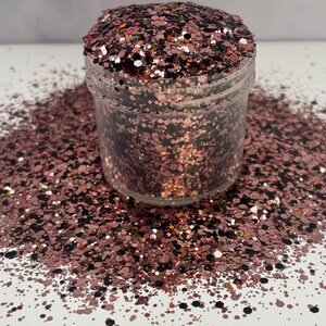 May include: A clear plastic jar filled with rose gold glitter, with some glitter spilled around the base. The glitter is a mix of shapes and sizes, including small dots and larger flakes, creating a sparkling effect.