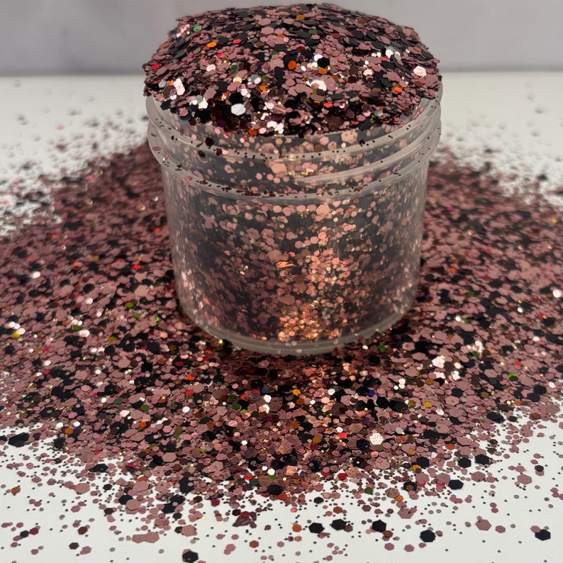 May include: A clear plastic jar filled with rose gold glitter, with some glitter spilled around the base. The glitter is a mix of shapes and sizes, including small dots and larger flakes, creating a sparkling effect.
