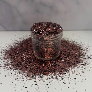 May include: A clear jar filled with mixed glitter in shades of brown, copper, black, and silver. The glitter is scattered around the jar on a white surface. The glitter is a mix of different shapes and sizes.
