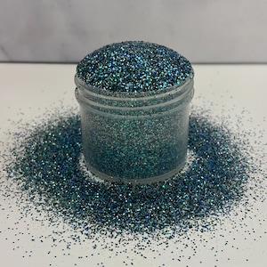 May include: A clear jar filled with teal and silver glitter. The glitter is scattered around the jar on a white surface.