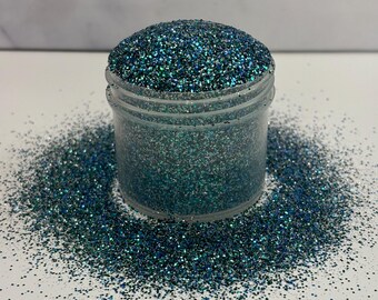 Op de afbeelding: Een glazen pot gevuld met teal en zilveren glitter. De glitter is rond de pot verspreid op een wit oppervlak.