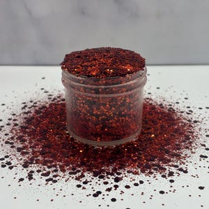 May include: A small glass jar filled with red glitter. The glitter is a mix of fine and chunky pieces, with a slight iridescent sheen. The jar is sitting on a white surface, and some glitter has spilled out around it.
