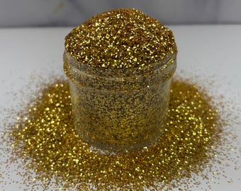 May include: A clear plastic jar filled with gold glitter, with some glitter spilled around the base. The glitter is a fine, reflective material, creating a sparkling effect. The jar sits on a white surface.