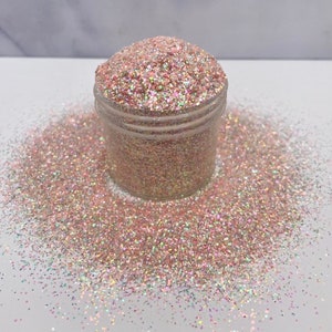 May include: A clear plastic jar filled with iridescent glitter in shades of pink, gold, and silver. The glitter is also scattered around the jar on a white surface. The glitter has a sparkling, reflective quality.