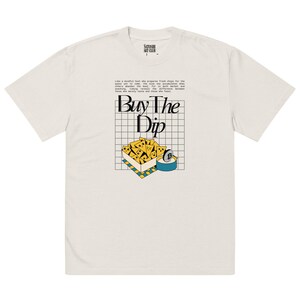 May include: A white t-shirt with a graphic of a box of yellow chips and a blue container with the text "Buy The Dip" in black.