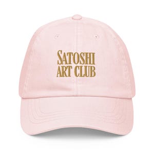Puede incluir: Una gorra de béisbol rosa con el texto "SATOSHI ART CLUB" bordado en dorado en la parte delantera.