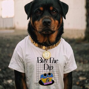 Pode incluir: Um cão Rottweiler vestindo uma camiseta branca com o texto "Buy The Dip" e um gráfico temático de Bitcoin. O cão também usa um colar de corrente dourada com um pingente de Bitcoin. O cão tem pelo preto e marrom.
