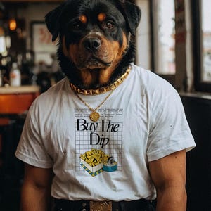 May include: A Rottweiler dog wearing a white t-shirt with a graphic print of a yellow box with the text "Buy The Dip" and a blue can. The dog is also wearing a gold chain necklace and a black belt with a gold buckle.