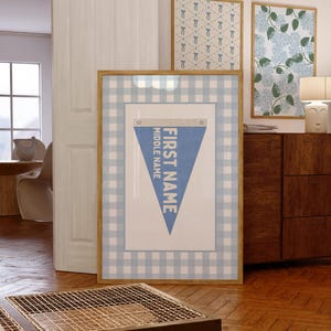 May include: A framed blue and white pennant with the words "FIRST NAME" and "MIDDLE NAME" on a light blue background. The frame has a light wood finish and is surrounded by a blue and white gingham pattern. Two other framed prints are visible in the background.