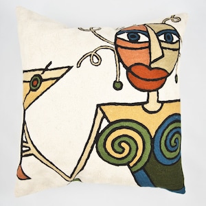 May include: White throw pillow with an embroidered design of a stylized woman with a martini glass. The woman has a colorful face with blue eyes, orange skin, and a red mouth. Her body is outlined in black and filled with yellow, green, and blue.