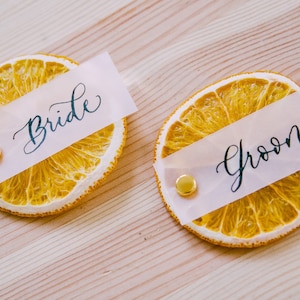 May include: Two dried orange slices with white paper place cards that say "Bride" and "Groom" in black calligraphy. The place cards are attached to the orange slices with gold tacks.
