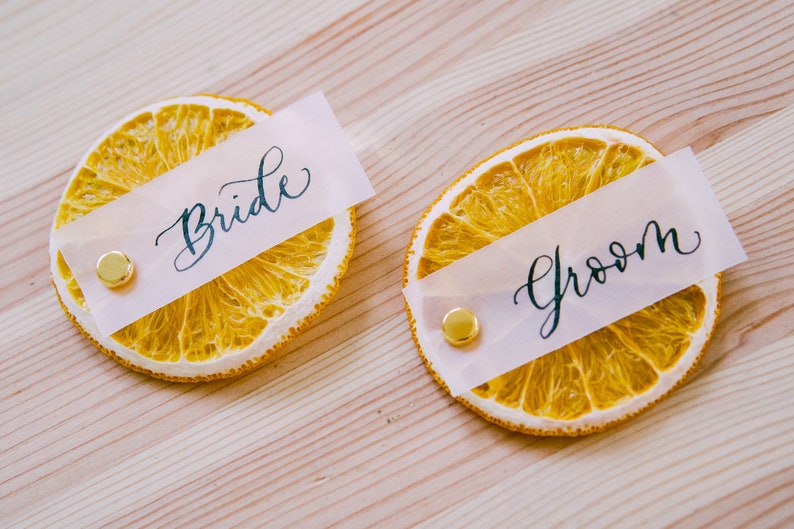 May include: Two dried orange slices with white paper place cards that say "Bride" and "Groom" in black calligraphy. The place cards are attached to the orange slices with gold tacks.