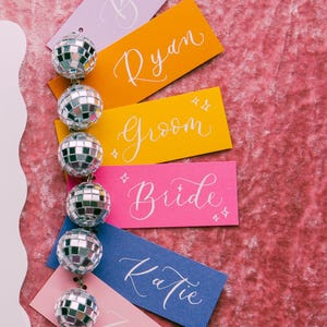 May include: A set of colorful place cards with names written in white calligraphy. The names are "Brian", "Ryan", "Groom", "Bride", "Katie", and "Zach". The place cards are decorated with silver disco balls.