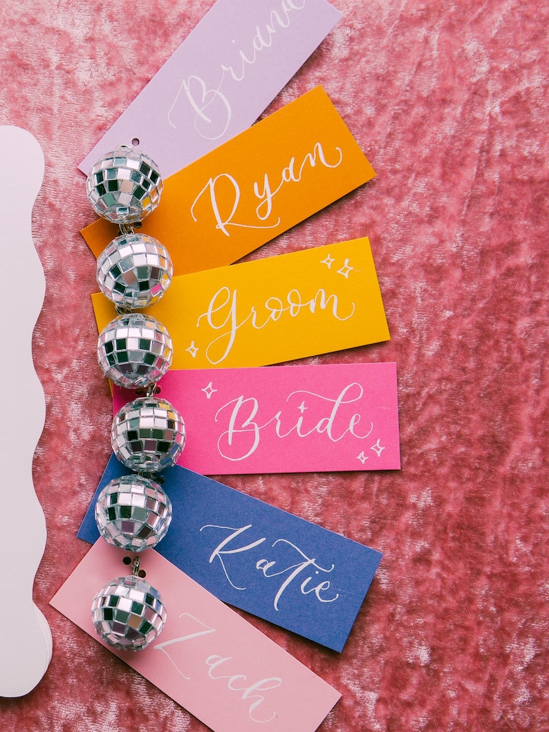 May include: A set of colorful place cards with names written in white calligraphy. The names are "Brian", "Ryan", "Groom", "Bride", "Katie", and "Zach". The place cards are decorated with silver disco balls.
