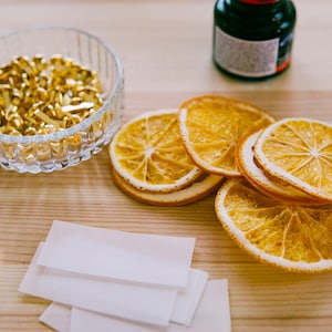 May include: A close-up of a craft project with dried orange slices, gold brads, white paper, and a black ink pen on a wooden surface.