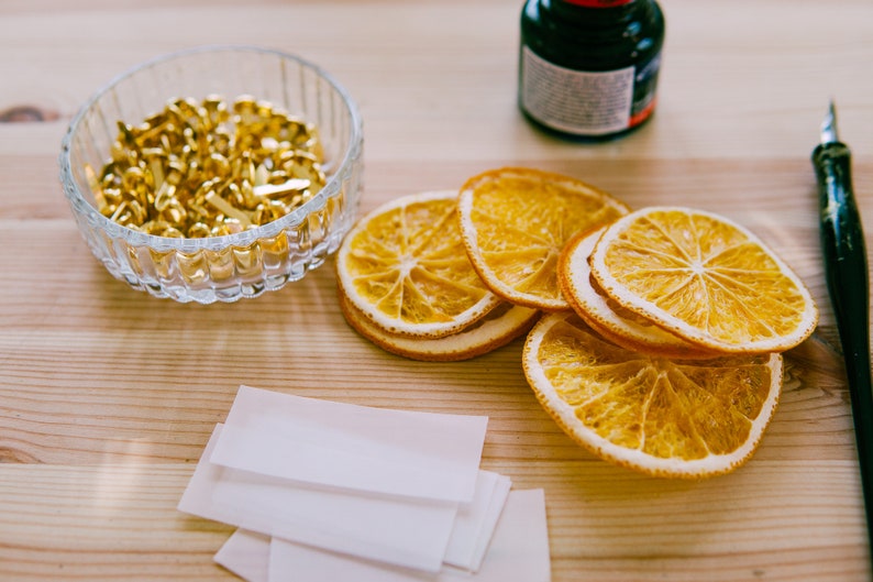 May include: A close-up of a craft project with dried orange slices, gold brads, white paper, and a black ink pen on a wooden surface.