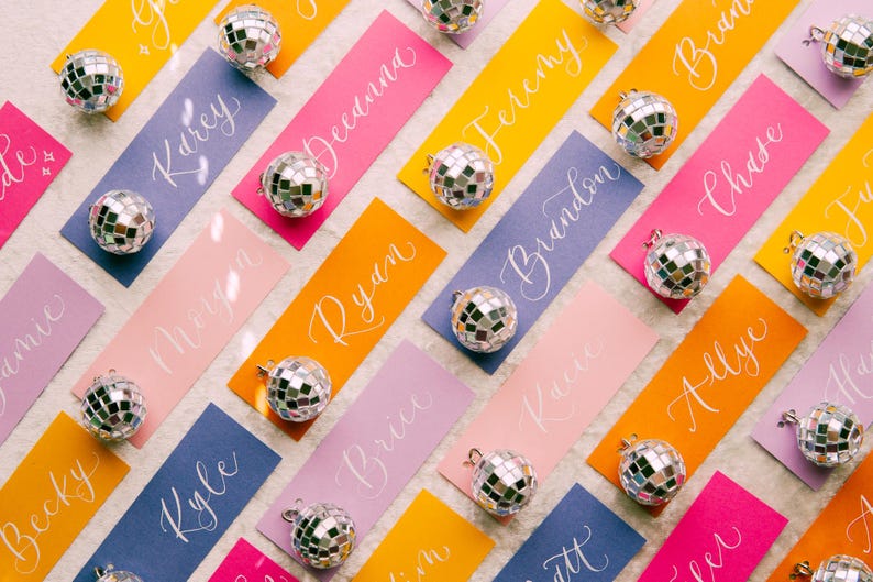May include: A collection of colorful place cards with white calligraphy names and silver disco ball ornaments. The names include: Amie, Kaley, Megan, Decanna, Kyle, Ryan, Jeremy, Brandon, Katie, Chase, Allyce, Becky, Brice, and more.