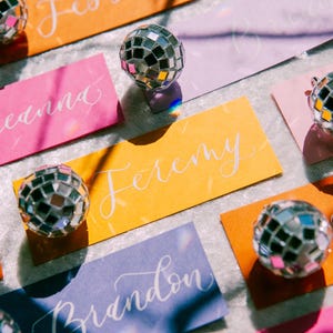 May include: Colorful place cards with names written in white calligraphy, each topped with a small silver disco ball. The place cards are on a white surface.