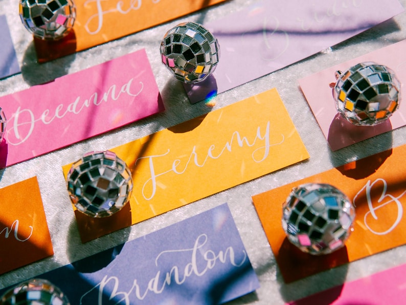 May include: Colorful place cards with names written in white calligraphy, each topped with a small silver disco ball. The place cards are on a white surface.