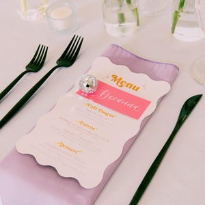 May include: A white menu with a pink and orange title card that says "Menu Deedana". The menu lists the courses: "First Course", "Entr&eacute;e", and "Dessert". The menu is on a purple napkin and a white tablecloth. There are two black forks and a black knife on the table.