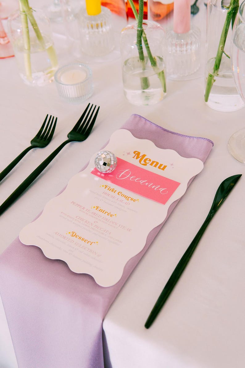 May include: A white menu with a pink and orange title card that says "Menu Deedana". The menu lists the courses: "First Course", "Entr&eacute;e", and "Dessert". The menu is on a purple napkin and a white tablecloth. There are two black forks and a black knife on the table.