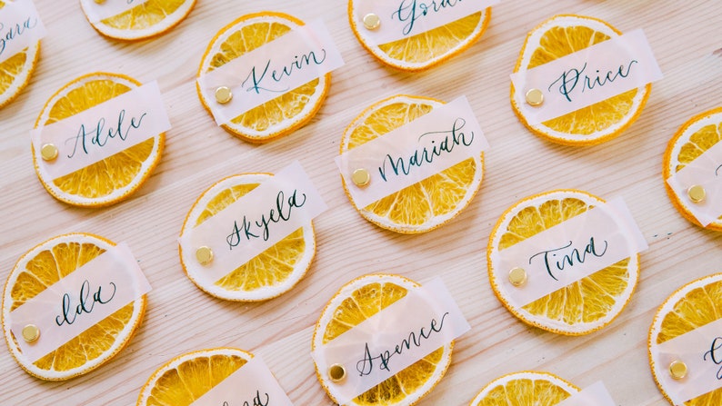 May include: Dried orange slices with white paper place cards with names written in calligraphy. The names include:  Addie, Lola, Kevin, Skyla, Mariah, Spence, Price, Tina.
