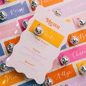 May include: A pink, white, and orange menu card with the word "Menu" in a retro font. The menu lists "First Course", "Entr&eacute;e", and "Dessert" with a disco ball charm attached to the card.