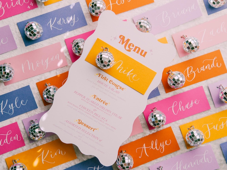 May include: A pink, white, and orange menu card with the word "Menu" in a retro font. The menu lists "First Course", "Entr&eacute;e", and "Dessert" with a disco ball charm attached to the card.